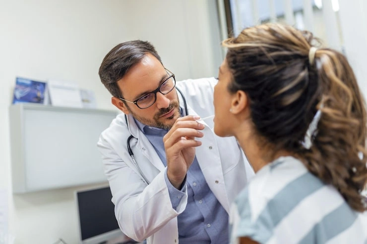 young woman sits exam table across from her doctor doctor reaches forward with tongue depressor as woman looks up sticks out her tongue 657921 823